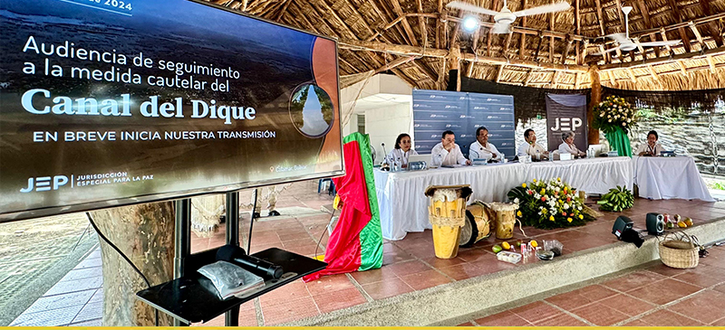 Foto de mesa de un panel dond e están 5 personas, con un televisor a un lado que dice audiencia de seguimiento a la medida cautelar del canal del dique
